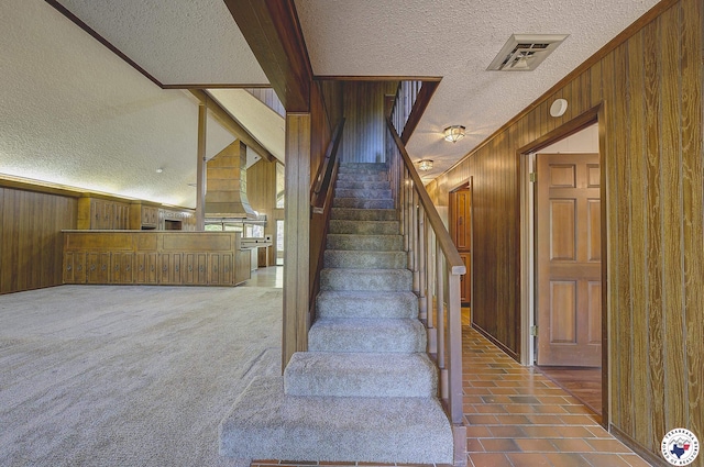 stairs with a textured ceiling, wood walls, ornamental molding, and visible vents