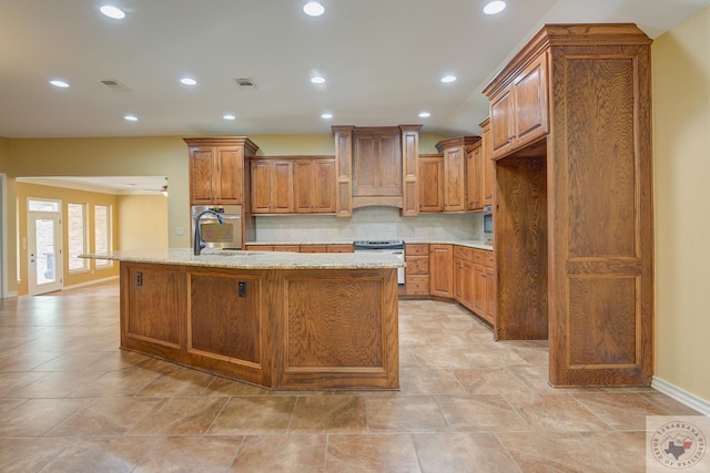 kitchen with light stone countertops, tasteful backsplash, sink, an island with sink, and stainless steel range