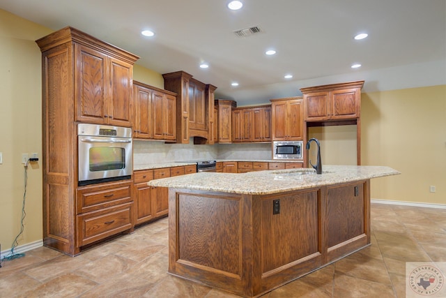 kitchen with appliances with stainless steel finishes, sink, light stone countertops, an island with sink, and decorative backsplash