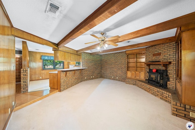 6514 Buchanan Loop, Texarkana, TX 75501 - unfurnished living room featuring a wood stove, wood walls, light carpet, beamed ceiling, and a ceiling fan