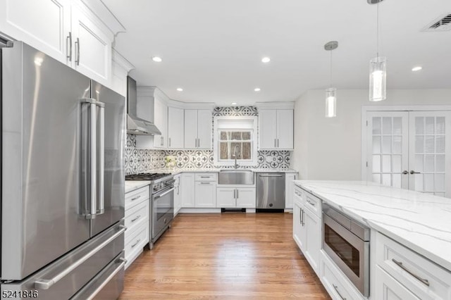 kitchen featuring high quality appliances, wall chimney range hood, hanging light fixtures, tasteful backsplash, and white cabinetry