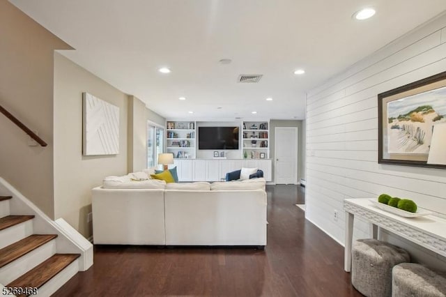 living area featuring built in shelves, dark wood-style flooring, visible vents, stairway, and wood walls