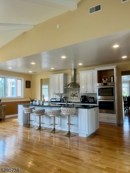 kitchen with a kitchen bar, stainless steel double oven, wall chimney exhaust hood, white cabinetry, and light wood-type flooring