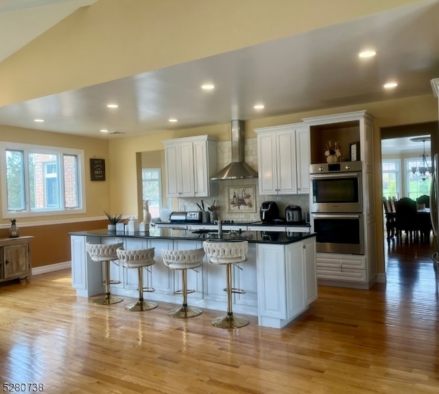 kitchen featuring wall chimney exhaust hood, a healthy amount of sunlight, light hardwood / wood-style flooring, a kitchen bar, and tasteful backsplash