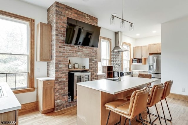 kitchen featuring a kitchen island with sink, wall chimney range hood, sink, hanging light fixtures, and light hardwood / wood-style flooring