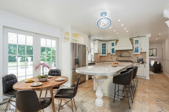 kitchen with custom exhaust hood, stainless steel fridge with ice dispenser, a kitchen island with sink, light tile patterned floors, and tasteful backsplash