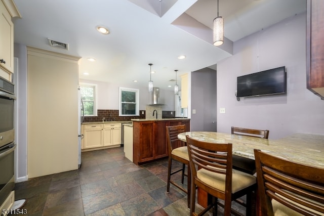 kitchen featuring decorative light fixtures, light stone counters, wall chimney range hood, tasteful backsplash, and a breakfast bar