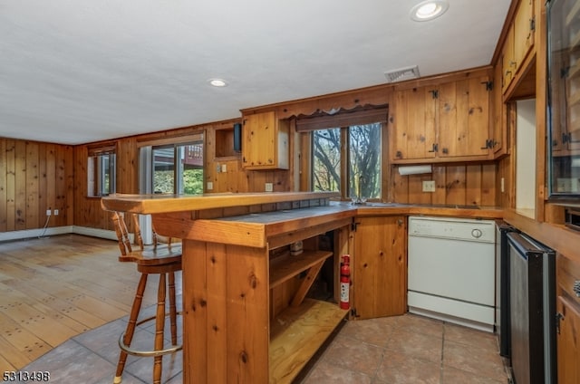 kitchen featuring light wood-type flooring, dishwasher, wood walls, kitchen peninsula, and a kitchen breakfast bar