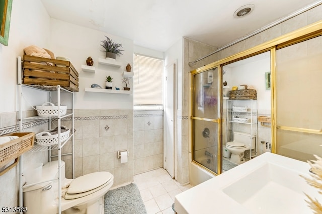 bathroom with tile walls, tile patterned flooring, combined bath / shower with glass door, and toilet