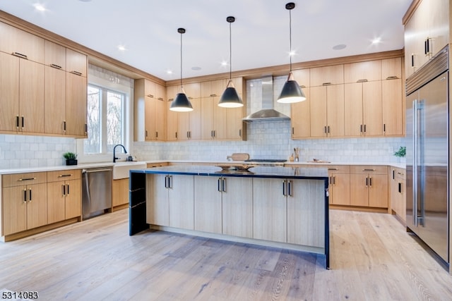 kitchen with light brown cabinets, stainless steel appliances, hanging light fixtures, and wall chimney range hood