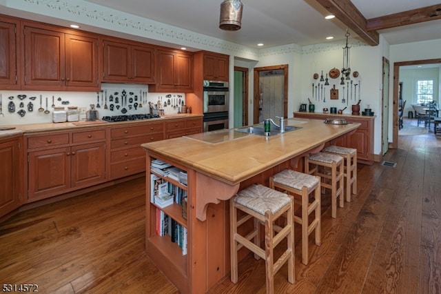 kitchen featuring a kitchen island with sink, beamed ceiling, dark wood-type flooring, and stainless steel double oven