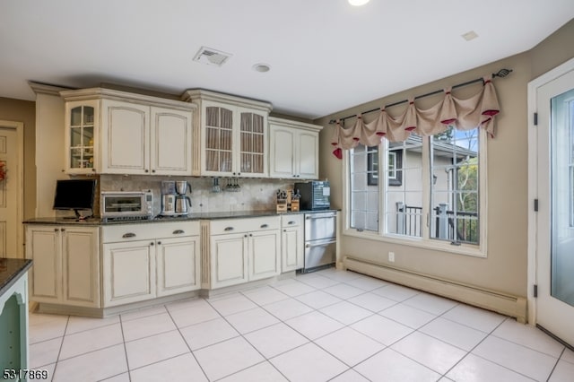 kitchen featuring cream cabinets, dark stone counters, a baseboard radiator, decorative backsplash, and light tile patterned floors