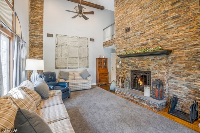 carpeted living room with a towering ceiling, ceiling fan, beam ceiling, and a stone fireplace