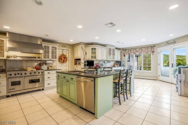 kitchen with wall chimney range hood, a kitchen breakfast bar, green cabinetry, stainless steel appliances, and an island with sink