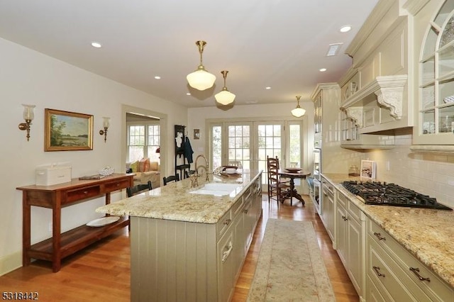 kitchen with a kitchen island with sink, a sink, gas cooktop, glass insert cabinets, and decorative backsplash