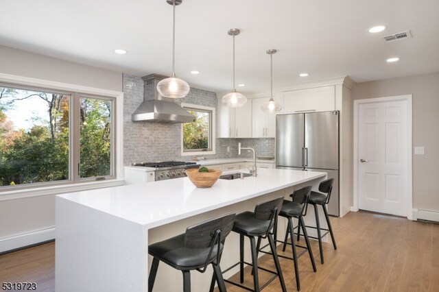 kitchen with sink, stainless steel appliances, wall chimney exhaust hood, hardwood / wood-style flooring, and a kitchen island with sink