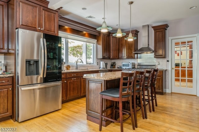 kitchen featuring a kitchen island, hanging light fixtures, stainless steel refrigerator with ice dispenser, light stone countertops, and wall chimney range hood