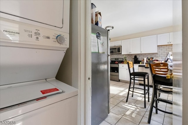 kitchen with appliances with stainless steel finishes, white cabinets, light tile patterned flooring, stacked washer / dryer, and tasteful backsplash