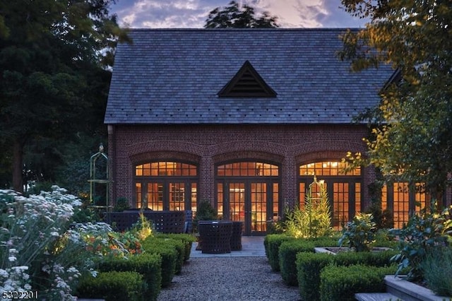 back of house at dusk featuring brick siding, a high end roof, and french doors