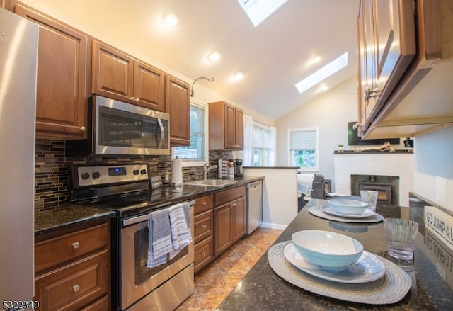 kitchen with appliances with stainless steel finishes, a brick fireplace, vaulted ceiling with skylight, dark stone countertops, and decorative backsplash