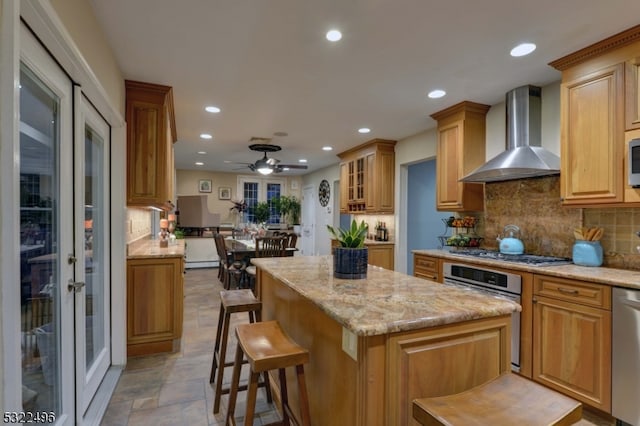 kitchen featuring wall chimney exhaust hood, a breakfast bar area, stainless steel appliances, light stone countertops, and ceiling fan