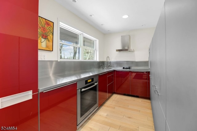 kitchen featuring oven, wall chimney range hood, stainless steel counters, light hardwood / wood-style flooring, and sink