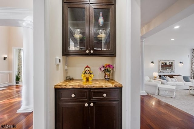 bar with dark wood-type flooring, ornate columns, crown molding, dark brown cabinets, and light stone countertops