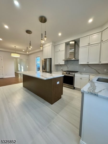 kitchen with appliances with stainless steel finishes, wall chimney exhaust hood, a kitchen island, white cabinetry, and hanging light fixtures