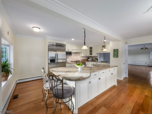 kitchen featuring stainless steel appliances, baseboard heating, a kitchen island with sink, pendant lighting, and white cabinets