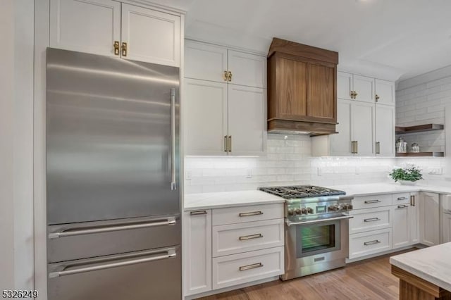 kitchen with premium range hood, decorative backsplash, light wood-type flooring, premium appliances, and white cabinetry