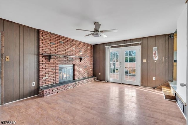 unfurnished living room featuring french doors, wooden walls, ceiling fan, light wood-type flooring, and a fireplace