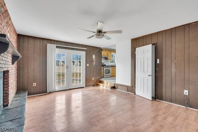 unfurnished living room with french doors, light wood-type flooring, a brick fireplace, ceiling fan, and wood walls