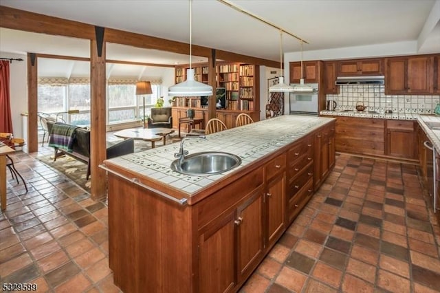 kitchen featuring backsplash, a kitchen island with sink, sink, pendant lighting, and tile counters