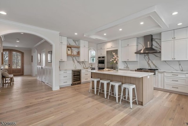 kitchen with open shelves, light countertops, wall chimney range hood, an island with sink, and beverage cooler