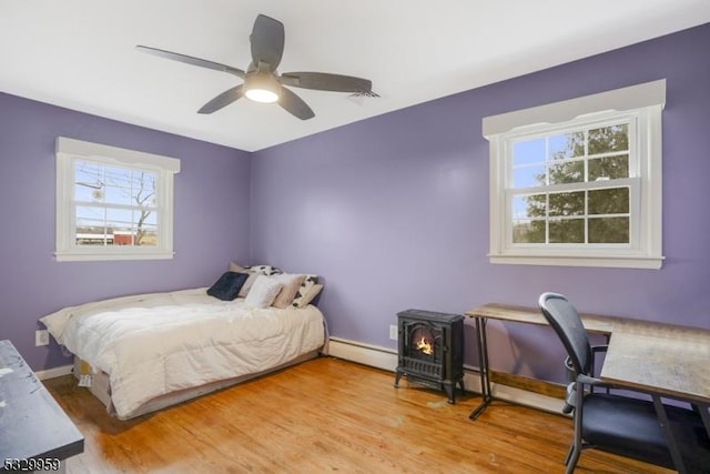 bedroom featuring wood-type flooring, baseboard heating, ceiling fan, and a wood stove