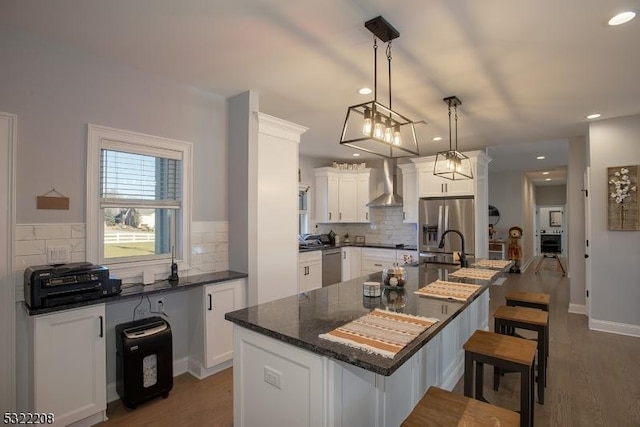 kitchen with white cabinets, decorative light fixtures, wall chimney range hood, and an island with sink