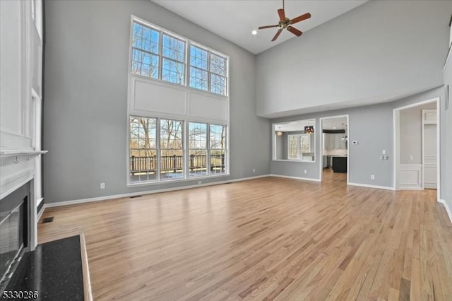 unfurnished living room featuring a high ceiling, a fireplace with flush hearth, ceiling fan, light wood-type flooring, and baseboards