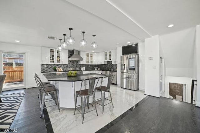 kitchen with visible vents, stainless steel refrigerator with ice dispenser, dark countertops, a kitchen breakfast bar, and wall chimney exhaust hood