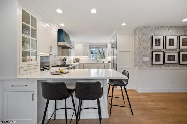 kitchen with a breakfast bar area, light hardwood / wood-style floors, white cabinets, kitchen peninsula, and wall chimney exhaust hood