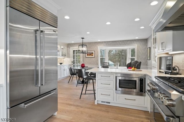 kitchen with pendant lighting, wall chimney range hood, white cabinetry, built in appliances, and light wood-type flooring