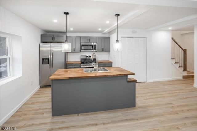 kitchen with stainless steel appliances, butcher block countertops, an island with sink, decorative light fixtures, and gray cabinets