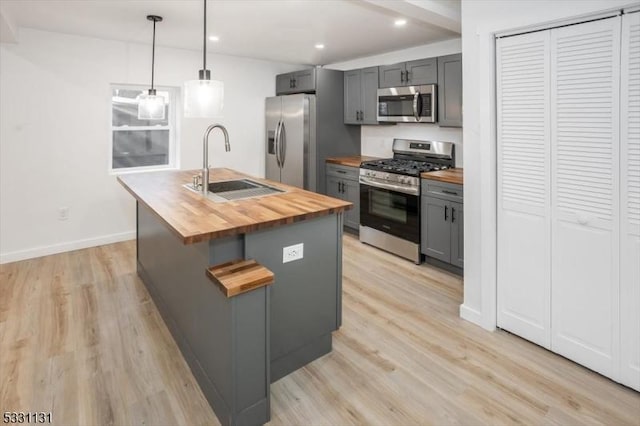 kitchen featuring butcher block counters, gray cabinetry, stainless steel appliances, an island with sink, and decorative light fixtures