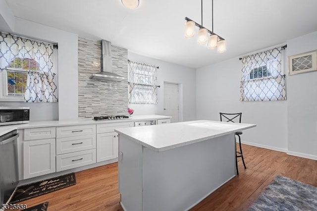 kitchen featuring a kitchen bar, wall chimney exhaust hood, stainless steel appliances, pendant lighting, and white cabinets