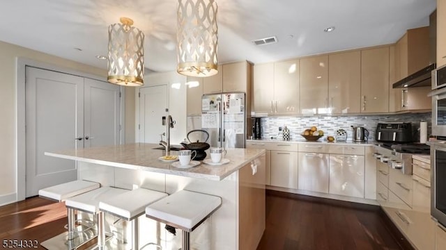 kitchen with backsplash, white refrigerator, decorative light fixtures, cream cabinetry, and an island with sink