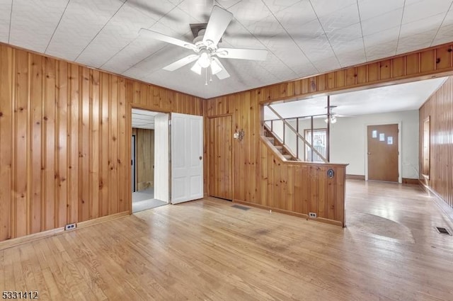 unfurnished room with light wood-style flooring, wood walls, baseboards, ceiling fan, and stairs