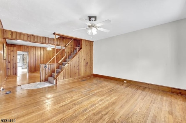 unfurnished living room with baseboards, stairway, wood walls, hardwood / wood-style floors, and a ceiling fan