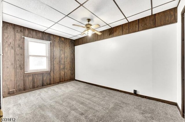 empty room featuring baseboards, ceiling fan, wood walls, carpet floors, and a paneled ceiling