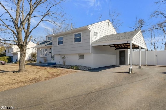 tri-level home featuring a carport, aphalt driveway, roof with shingles, and fence