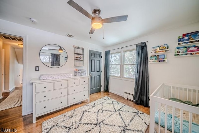 bedroom featuring ceiling fan, light hardwood / wood-style flooring, a nursery area, and a baseboard heating unit