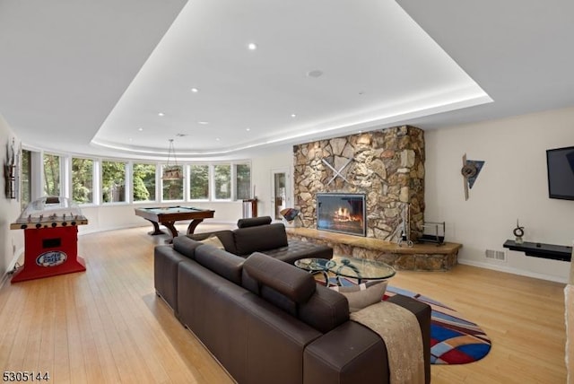 living room with a tray ceiling, billiards, light hardwood / wood-style flooring, and a stone fireplace